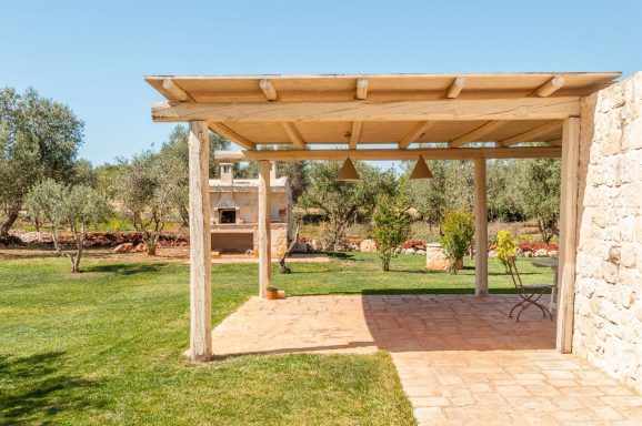 Patio with a wooden pergola, olive trees, and a stone outdoor fireplace in a sunny garden.