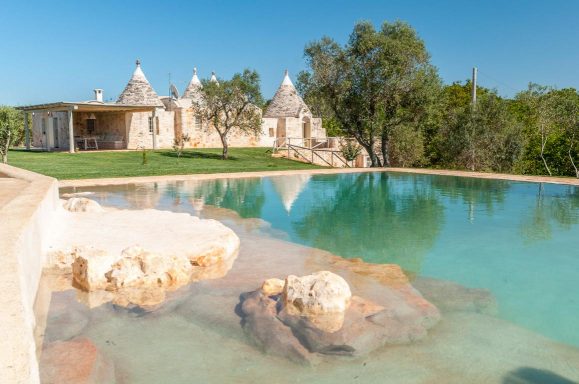 Trullo house with a swimming pool and lush greenery under a clear blue sky.